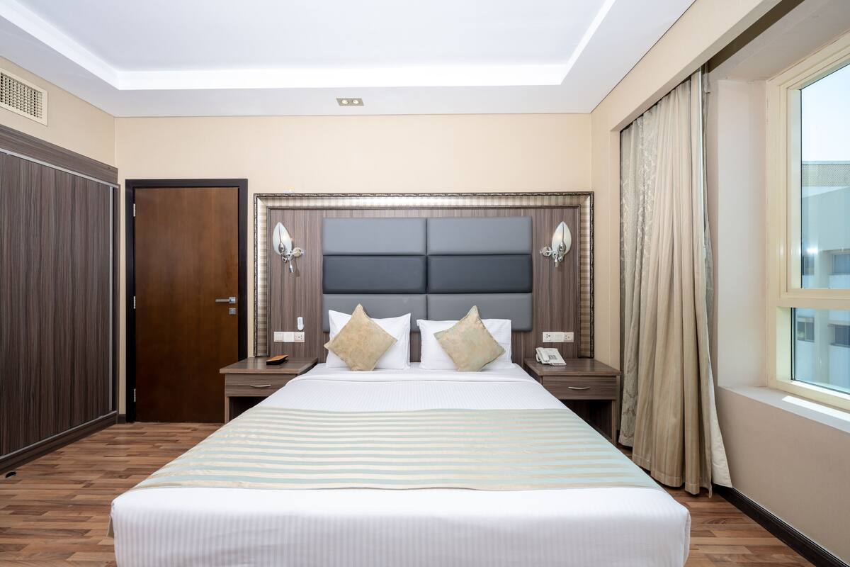 A hotel room with a large bed, a wooden wardrobe, a nightstand with a telephone, and a window with curtains.