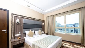 Ein Hotelzimmer mit einem großen Bett, einem Nachttisch, einem Holz-Kopfteil, einem Fenster mit Blick auf die Stadt und einer Tür.