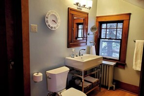 Un baño con inodoro blanco, lavamanos con espejo superior, un reloj en la pared y una ventana con vista a árboles.