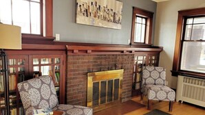 Un salon chaleureux avec une cheminée, deux fauteuils à motifs, une bibliothèque et un grand tableau abstrait accroché au mur.