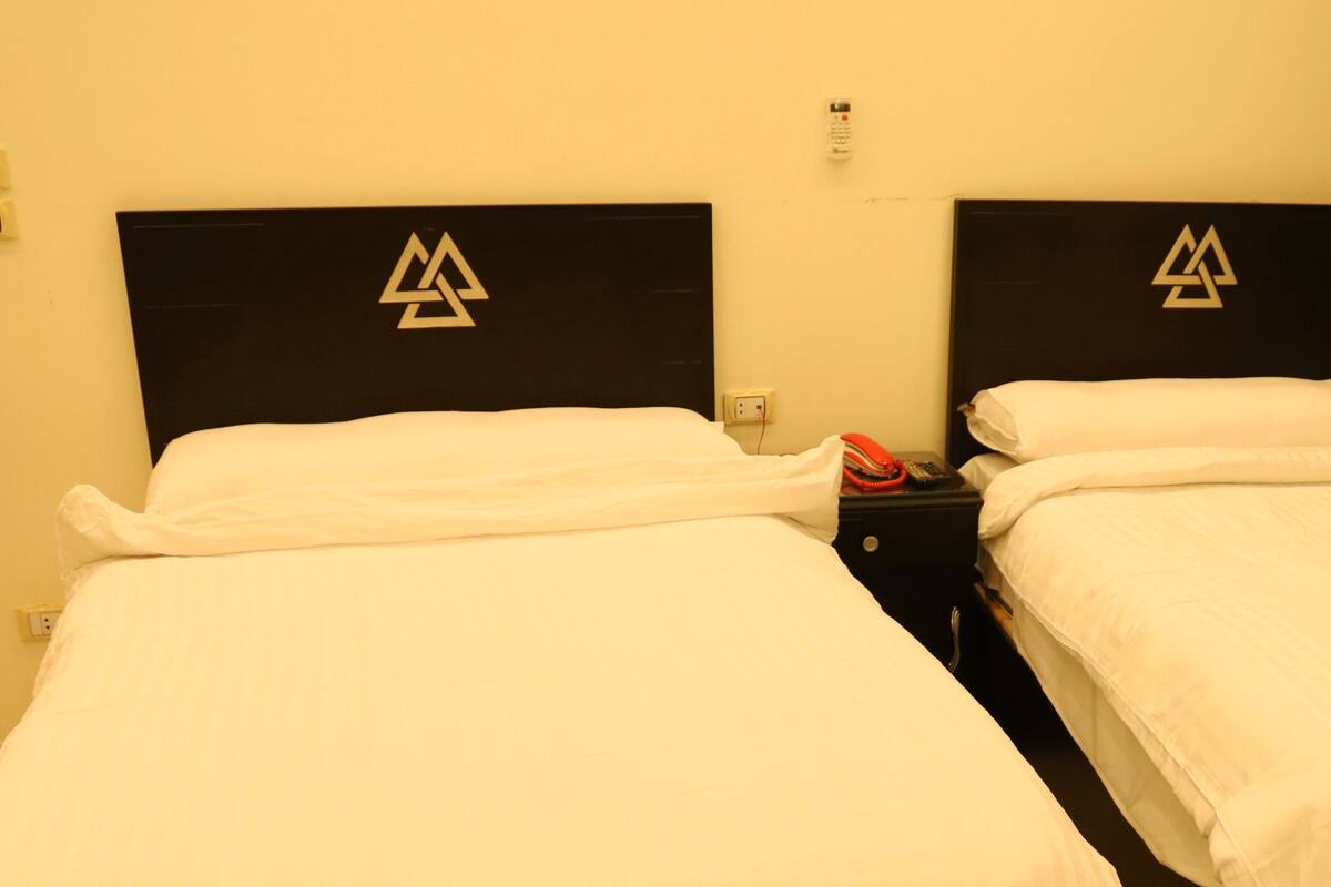 A hotel room with two beds, each with a black headboard featuring a logo, white bedding, and a nightstand with a phone and charger.