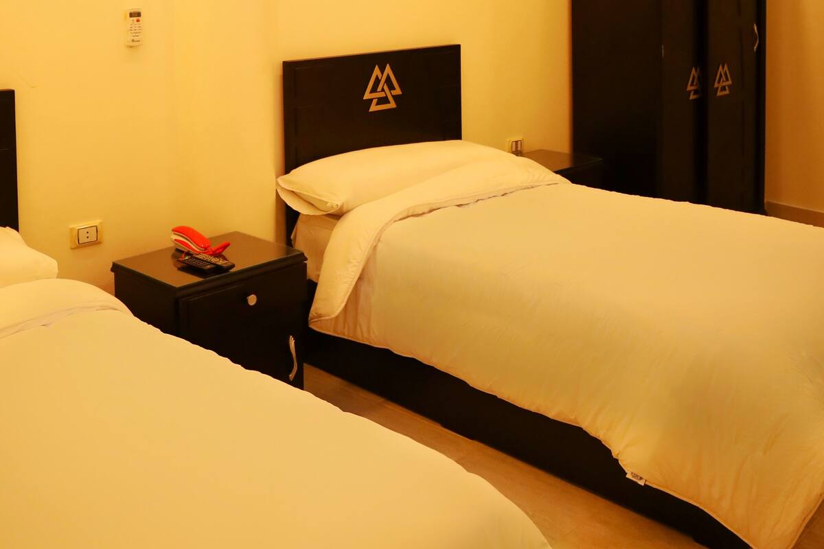 A hotel room with two beds, a black wardrobe, a bedside table with a phone and a red object, and a wall-mounted control panel.