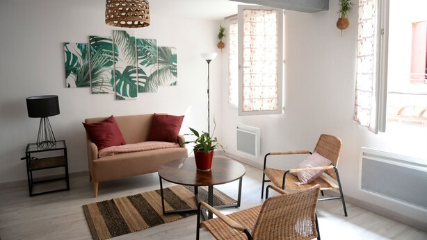 A modern living room with a sofa, two wicker chairs, a coffee table, and a floor lamp. There are large tropical leaf paintings on the wall, a window with curtains, and a hanging plant. - Large studio near the ancient theater in Orange (Orange)