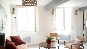 A modern living room with a sofa, two wicker chairs, a round coffee table, and a hanging light fixture. There are two windows with curtains, and a potted plant on the windowsill. - Large studio near the ancient theater in Orange (Orange)