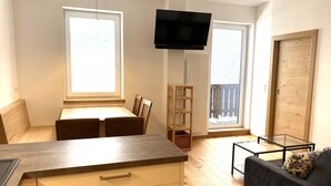 Kompakti oleskelutila, jossa on pieni keittiö, ruokailualue, sohva, seinälle asennettu televisio ja parveke, jossa on puinen kaide.