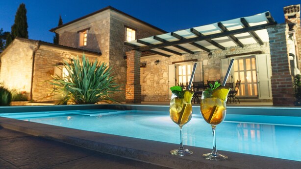 Vista de la piscina con dos bebidas, un edificio de piedra y una pérgola.
