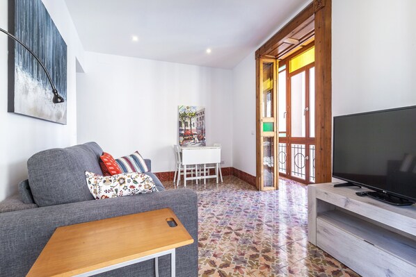 A modern living room with a grey sofa, a wooden coffee table, a TV on a wooden stand, a white desk, and a colorful tiled floor.