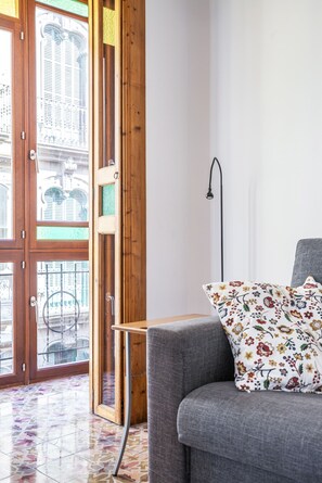 A room with a grey sofa, a floral pillow, a floor lamp, and a view of a building through a wooden door.