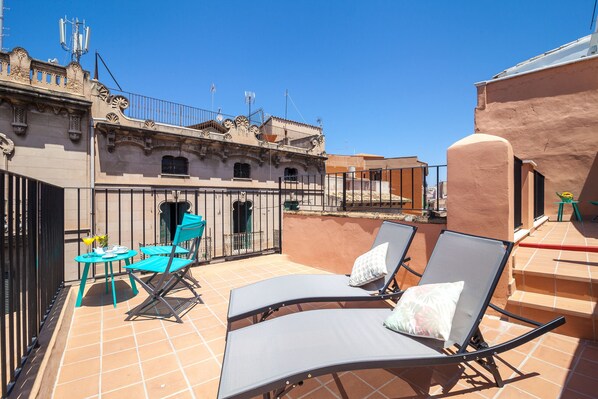 A rooftop terrace with two lounge chairs and a table, surrounded by a wrought iron fence and a building with ornate architecture.