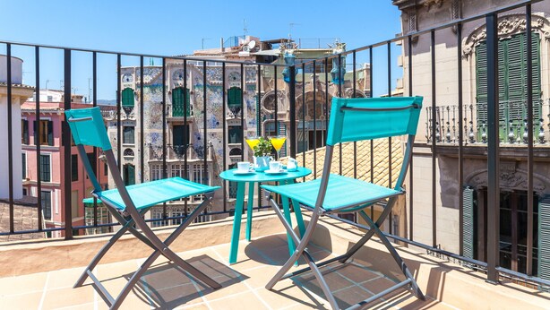 A balcony with turquoise chairs and a small table, overlooking a cityscape with historic buildings.