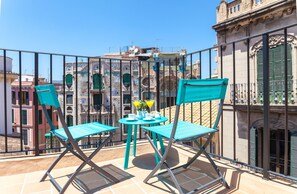 Un balcón con sillas turquesas y una pequeña mesa, con vistas a un paisaje urbano con edificios históricos.