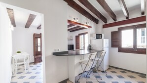 Una cocina con encimera blanca, microondas y refrigerador. Hay dos sillas y una mesa con una planta. El piso tiene un patrón de cuadros.