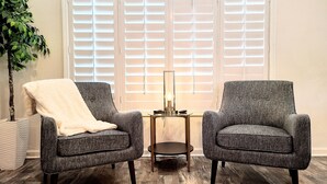 Two grey armchairs with a white throw blanket, a small table with a lamp, and a large window with blinds.