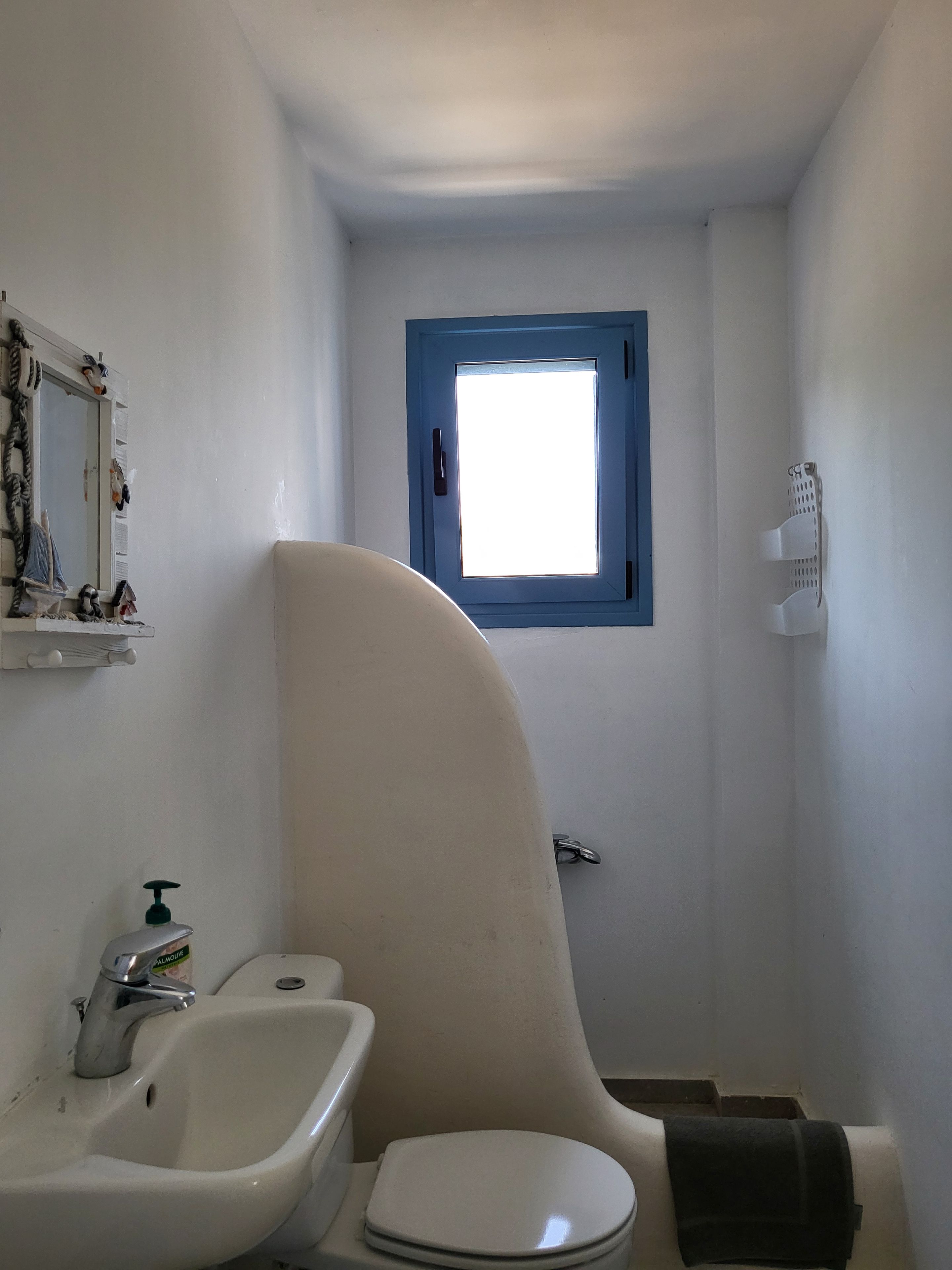 Ein kleines Badezimmer mit einem weißen Waschbecken, einer Toilette, einem Spiegel und einem kleinen Fenster mit blauem Rahmen.