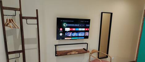 Habitación de hotel con una cama, una silla de madera, un televisor montado en la pared mostrando diferentes servicios de streaming, un estante de madera debajo del televisor y una escalera de madera apoyada contra la pared.