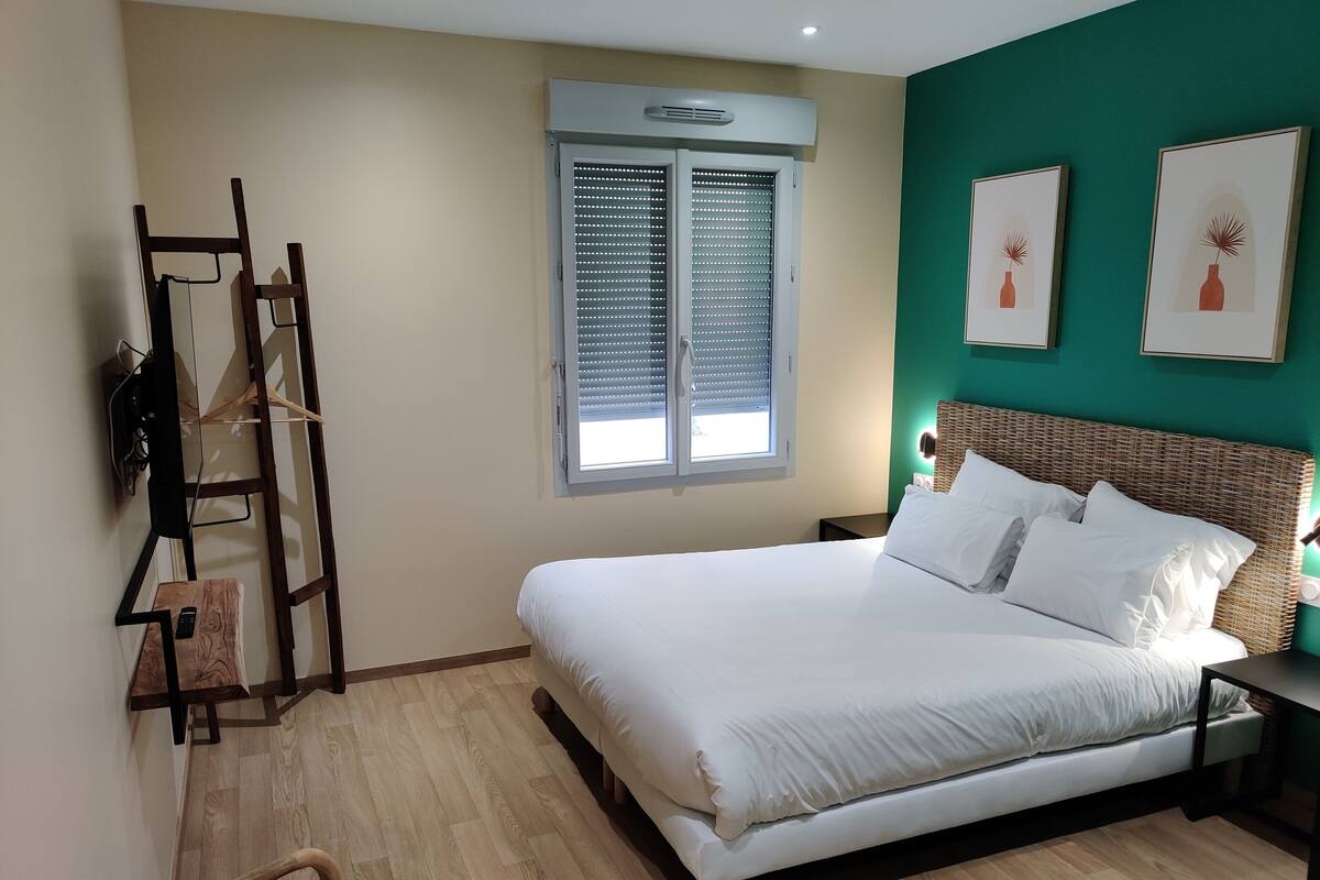 Une chambre à coucher comprenant un lit, une fenêtre avec des stores, un téléviseur fixé au mur, une table de chevet et une chaise.