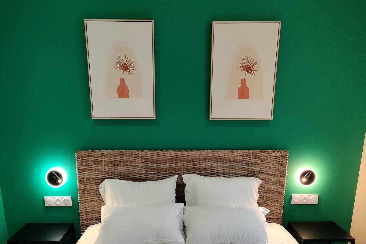 Un lit bien fait, avec du linge de lit blanc et deux serviettes pliées posées sur le lit. On y trouve deux tables de chevet avec des lampes fixées au mur et une tête de lit en osier.