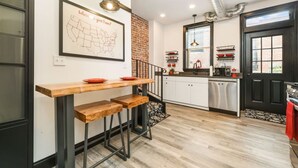 A modern kitchen with a wooden table and stools, a map on the wall, and a black door. - Brand New ★ Prime Location ★ Exposed Brick Beauty! (Pittsburgh)