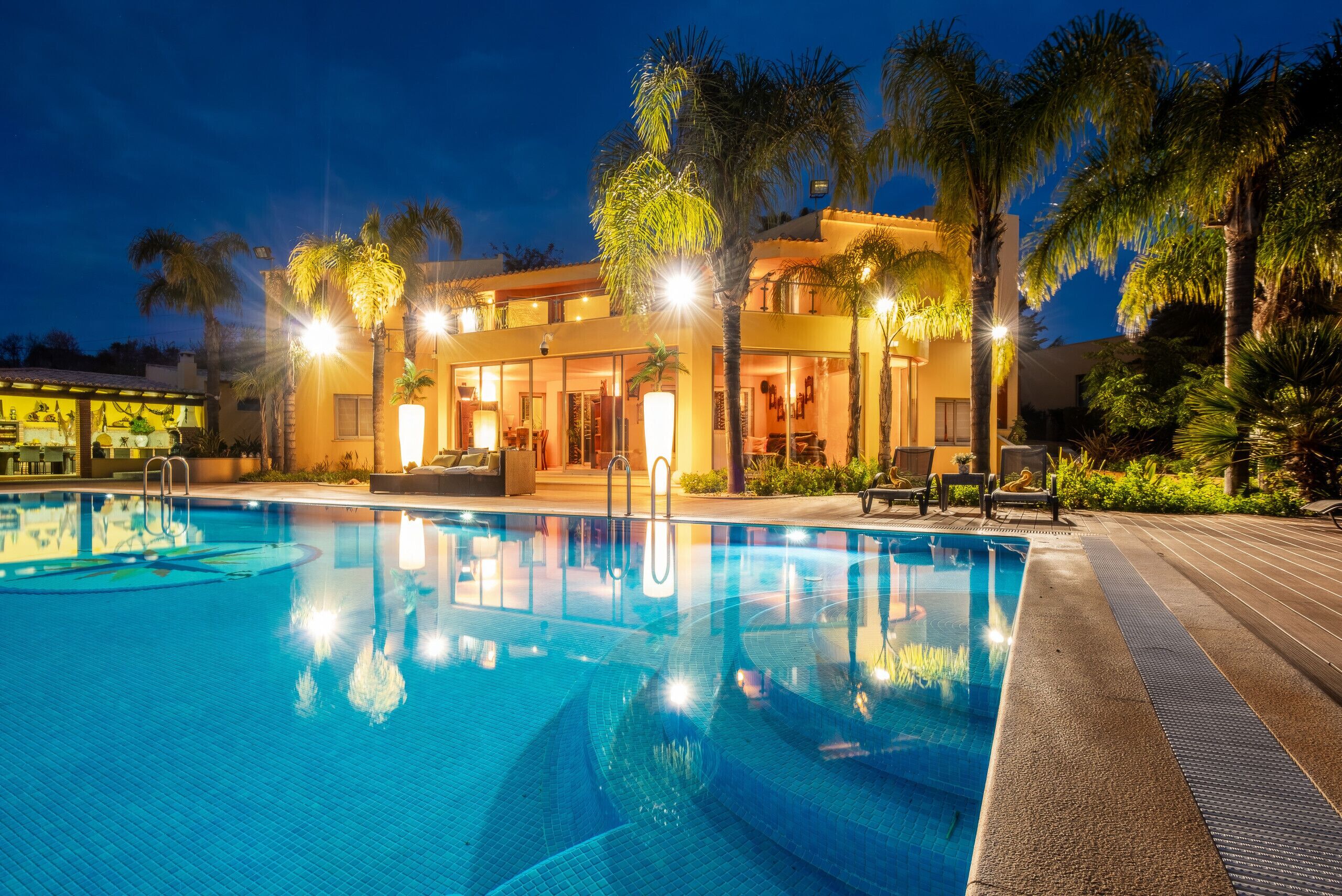 A swimming pool with a clear blue water surface, surrounded by palm trees and a building with multiple levels and balconies.