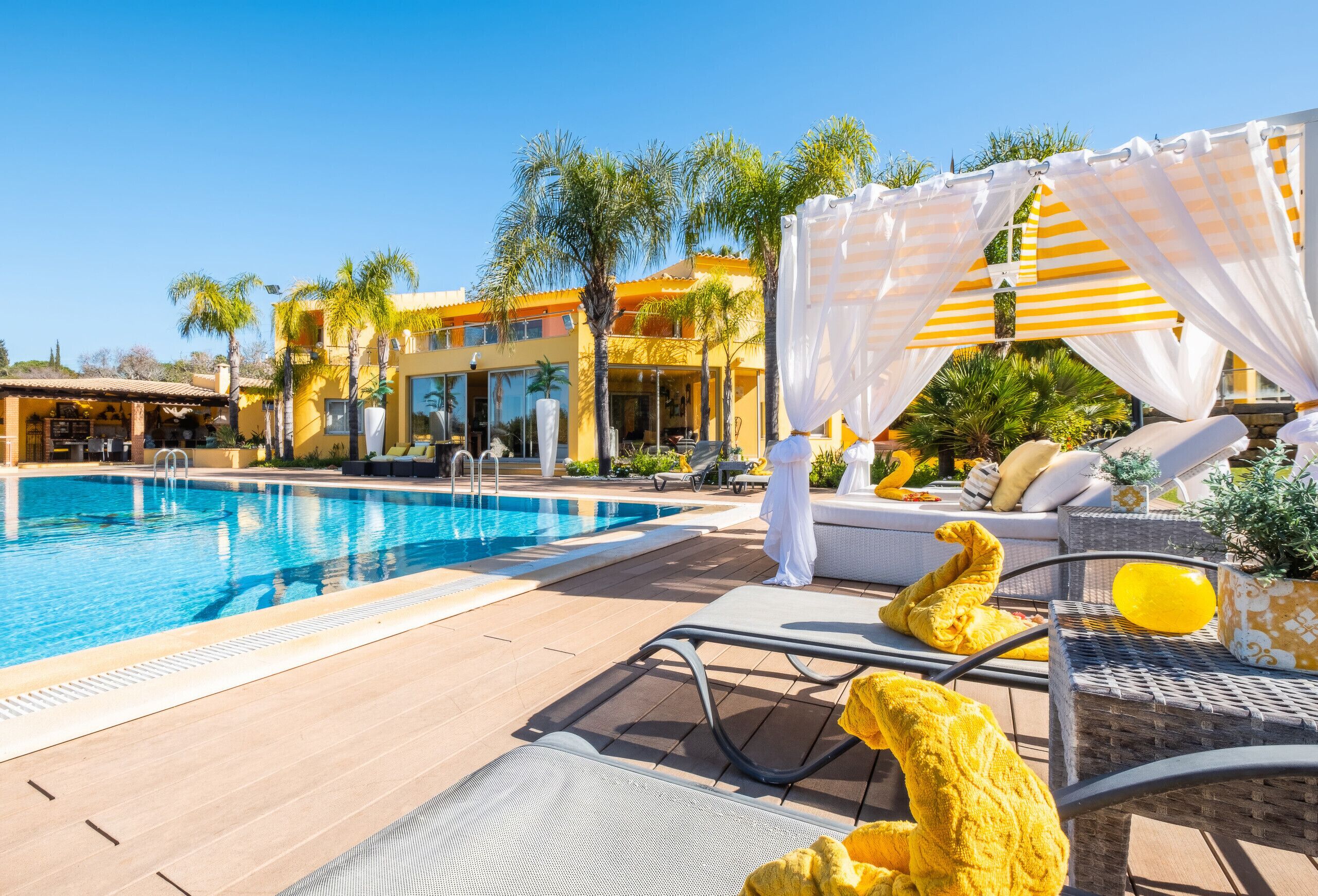 A poolside area with a clear blue pool, lounge chairs, and a building with balconies in the background.