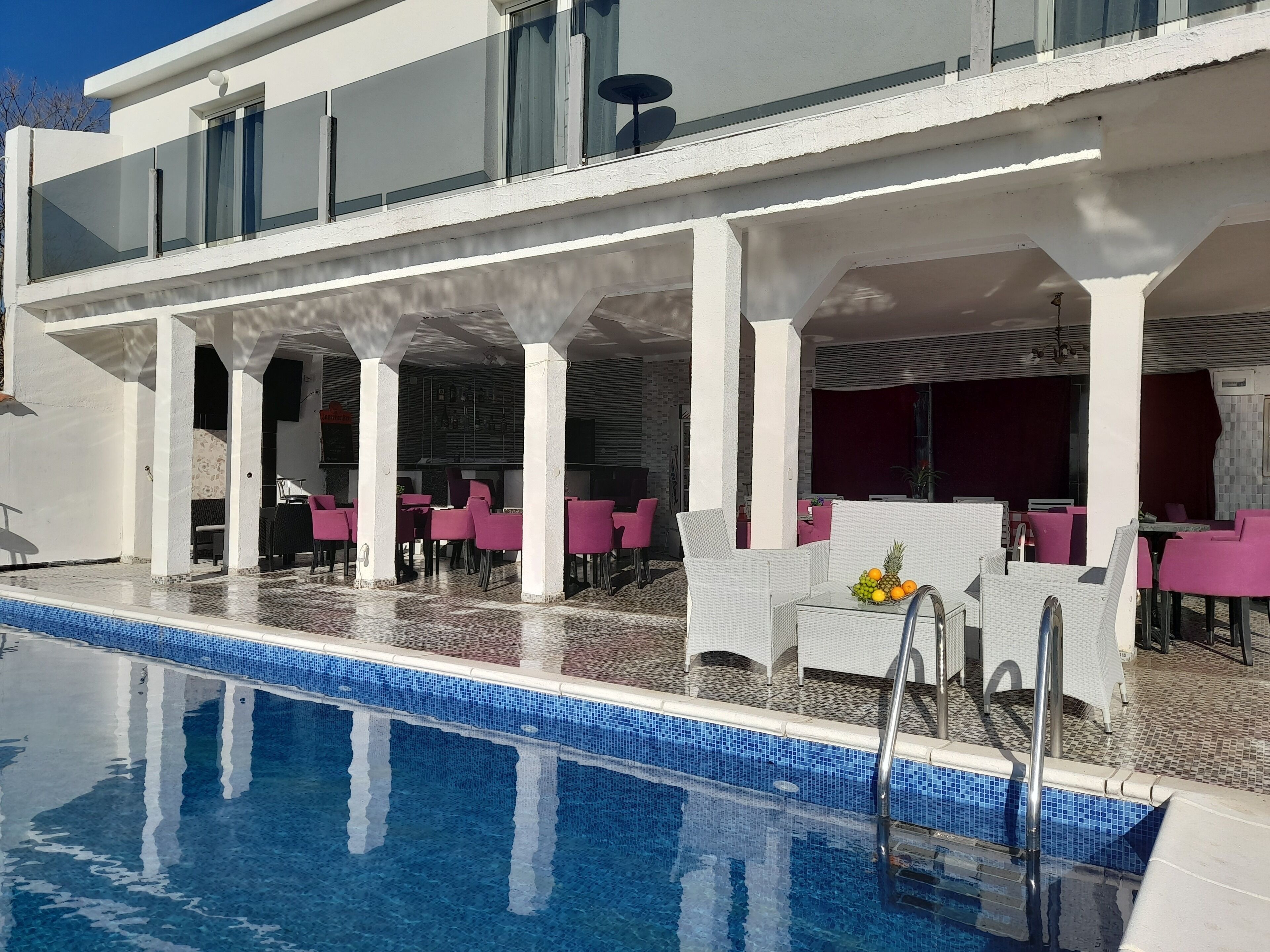 A poolside area with a clear blue pool, white pillars, and pink chairs.