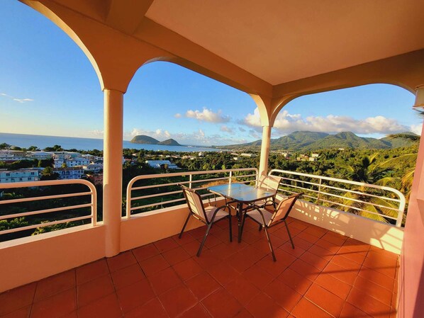 A balcony with a table and chairs, offering a view of a coastal town and mountains.