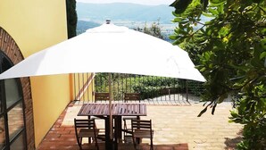 Un patio con mesa y sillas bajo una sombrilla blanca, con vistas a un paisaje pintoresco.