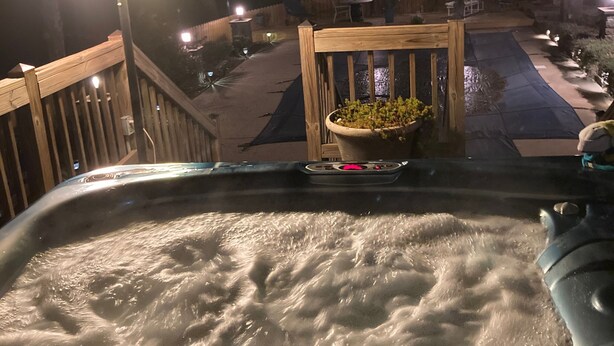 Jacuzzi con cubierta de madera y mobiliario exterior, rodeado por una cerca de madera e iluminado con luces para exteriores.