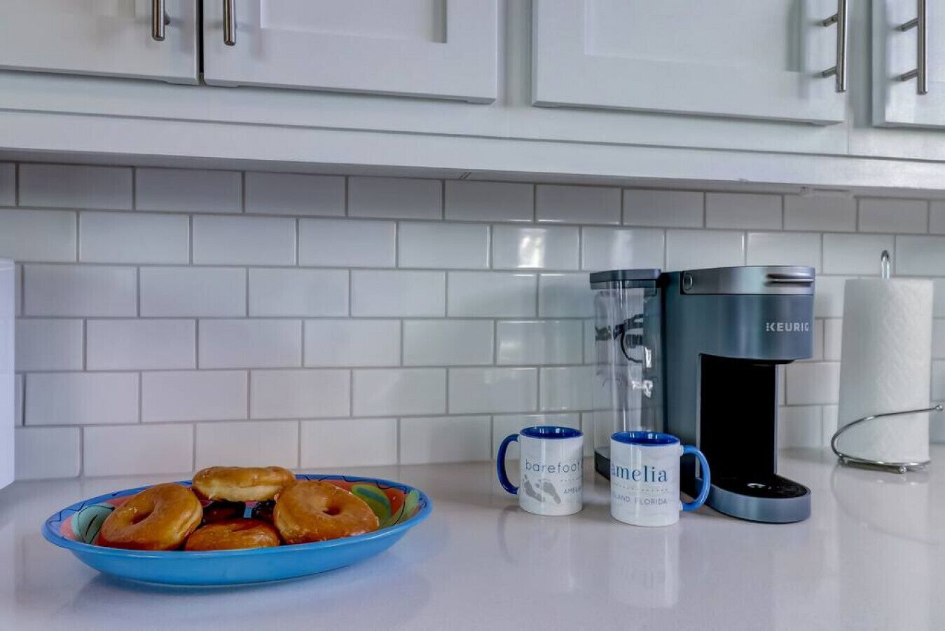 Une cuisine avec des meubles blancs, une machine à café, deux tasses et un bol rempli de beignets.