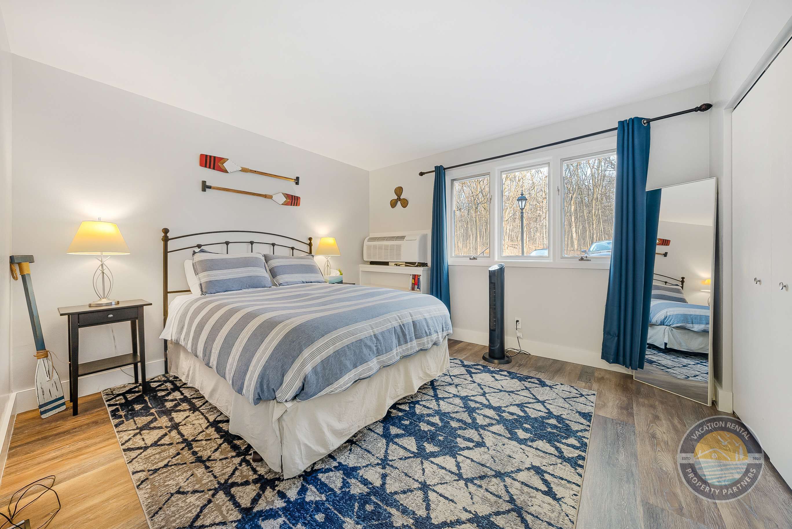 A bedroom with a bed, bedside tables, a window with curtains, a mirror, and a patterned rug.
