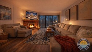 A cozy living room with a fireplace, comfortable seating, and a large window offering a view of trees. - Lake Geneva Condo with Pool, Fireplace & Parking (Lake Geneva)