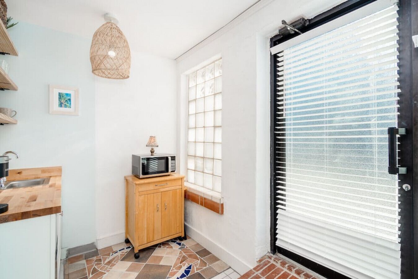 Una cocina compacta con microondas sobre un mueble de madera, un lavamanos pequeño y una ventana con persianas.