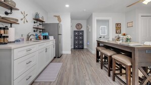 Une cuisine moderne avec des meubles blancs, un plan de travail en bois et un coin bar avec des tabourets. On y trouve un réfrigérateur, des étagères remplies d’ustensiles de cuisine et un ventilateur de plafond.