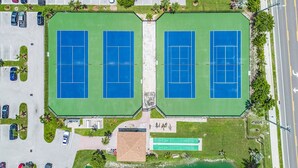 Vue aérienne d’un complexe sportif comprenant deux courts de tennis, des aires de stationnement et un petit bâtiment sur lequel on peut voir une enseigne.