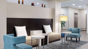 A modern hotel lobby with a sofa, two armchairs, a small table, a lamp, and a ceiling-mounted shelf with decorative items. - La Quinta Inn & Suites by Wyndham Wenatchee (Wenatchee)
