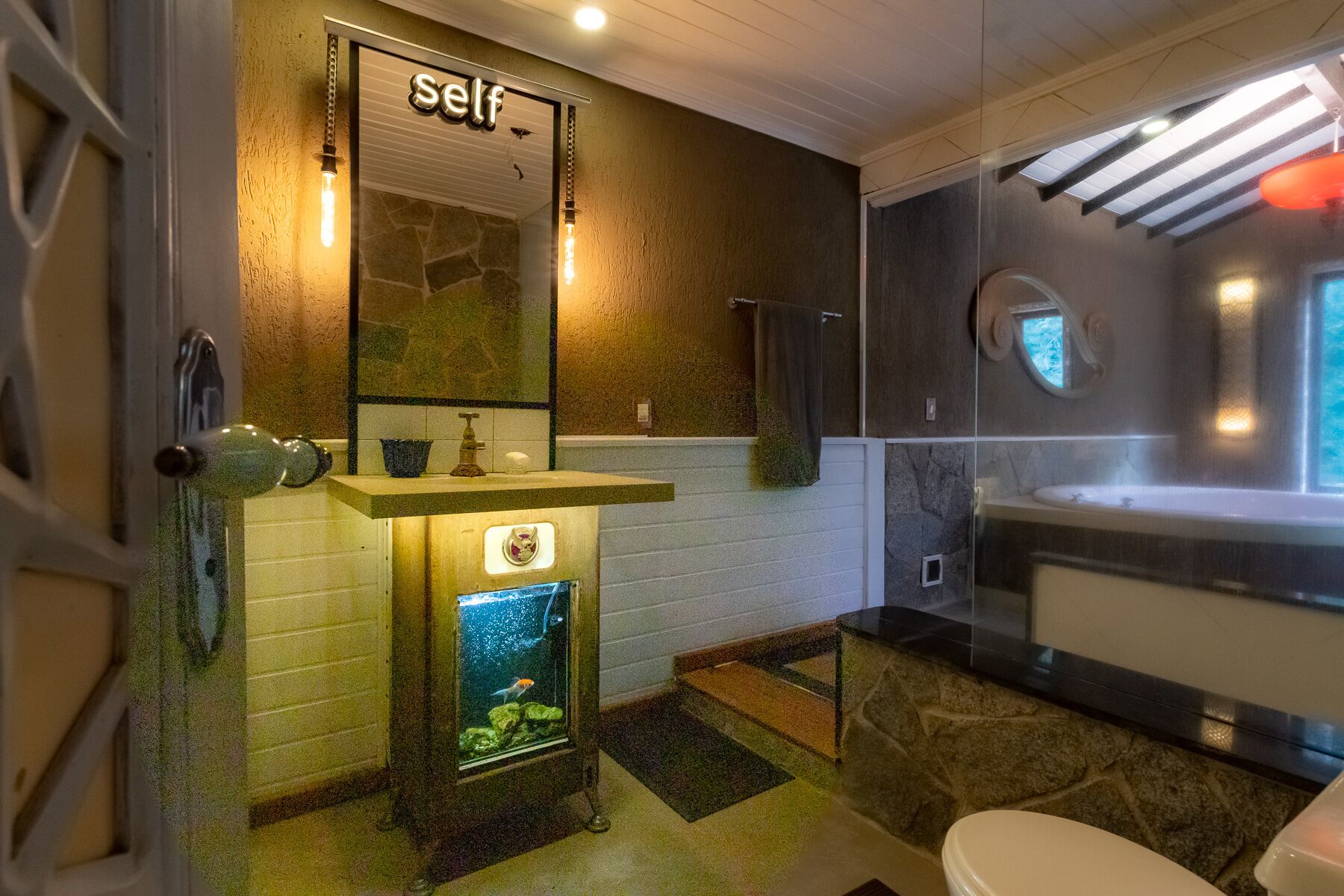 Ein Badezimmer mit Steinwand, Waschbecken mit Spiegel, Toilette und einem kleinen Aquarium.