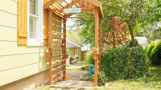 Une pergola en bois surmontée d’une enseigne indiquant « JARDIN », un chemin menant à une maison et un jardin luxuriant agrémenté de diverses plantes et fleurs.