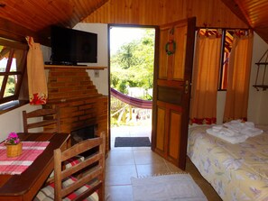 A cozy cabin interior with a wooden staircase, a TV, a bed with floral bedding, and a view of a hammock outside. - pousada caminho do escorrega (Itatiaia)