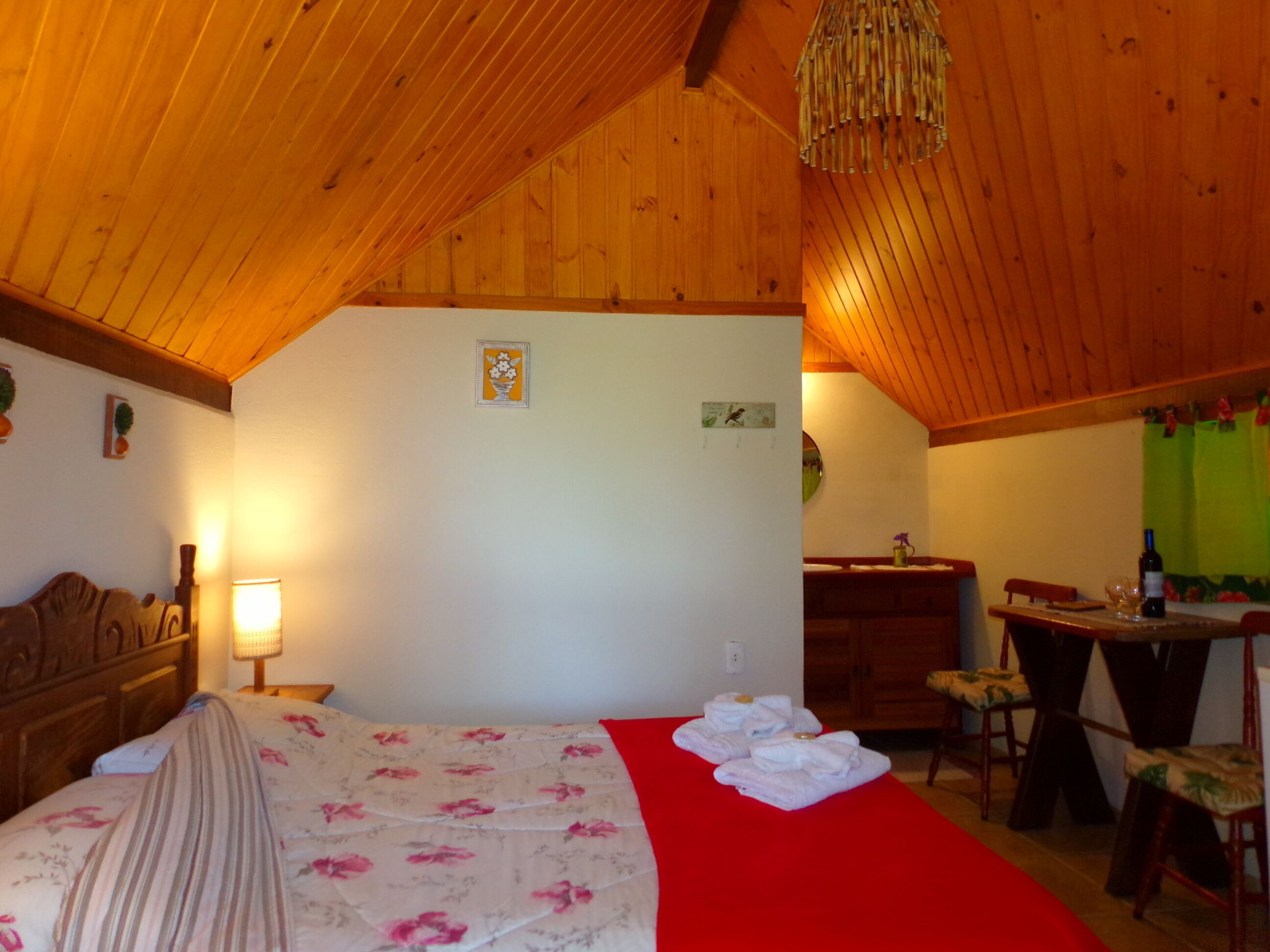 Una camera accogliente con soffitto in legno, un letto con biancheria da letto floreale, una coperta rossa, una scrivania con una sedia e un piccolo tavolino con una bottiglia.