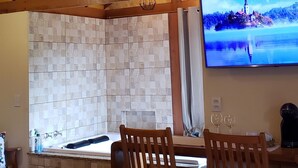 A cozy room with a wooden ceiling, a stone wall, a bed with white bedding, two wooden chairs, and a TV displaying a scenic view. - Pousada Caminho do Escorrega (Itatiaia)