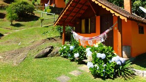 A small, single-story house with a red roof and orange walls, surrounded by lush greenery and a hammock. - Pousada Caminho do Escorrega (Itatiaia)
