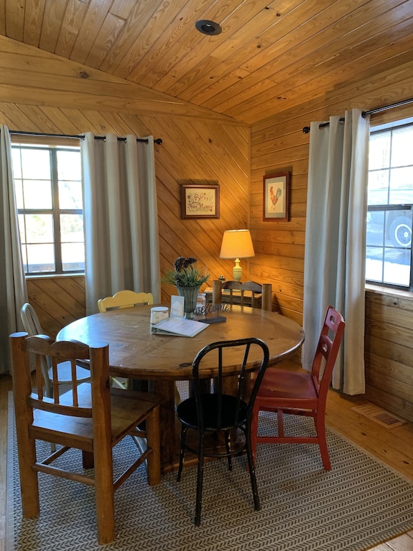 A wooden-paneled room with a round wooden table, four chairs, a lamp, and two windows with curtains.