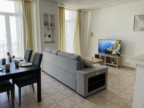 Un salon moderne avec un canapé gris, une table à manger avec des chaises, un téléviseur posé sur un meuble et un balcon dot é d’une rambarde.