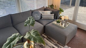 A modern living room with a grey sectional sofa, a small side table with candles and a plant, and a large window with blinds. - Remodeled Colonial Retreat Close to  Shops and Dining (Wichita)