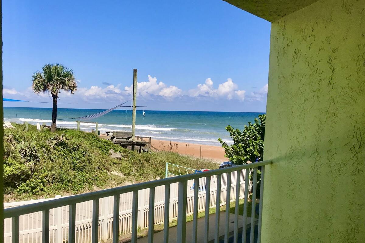 A balcony with a view of a beach, a hammock, and a palm tree.