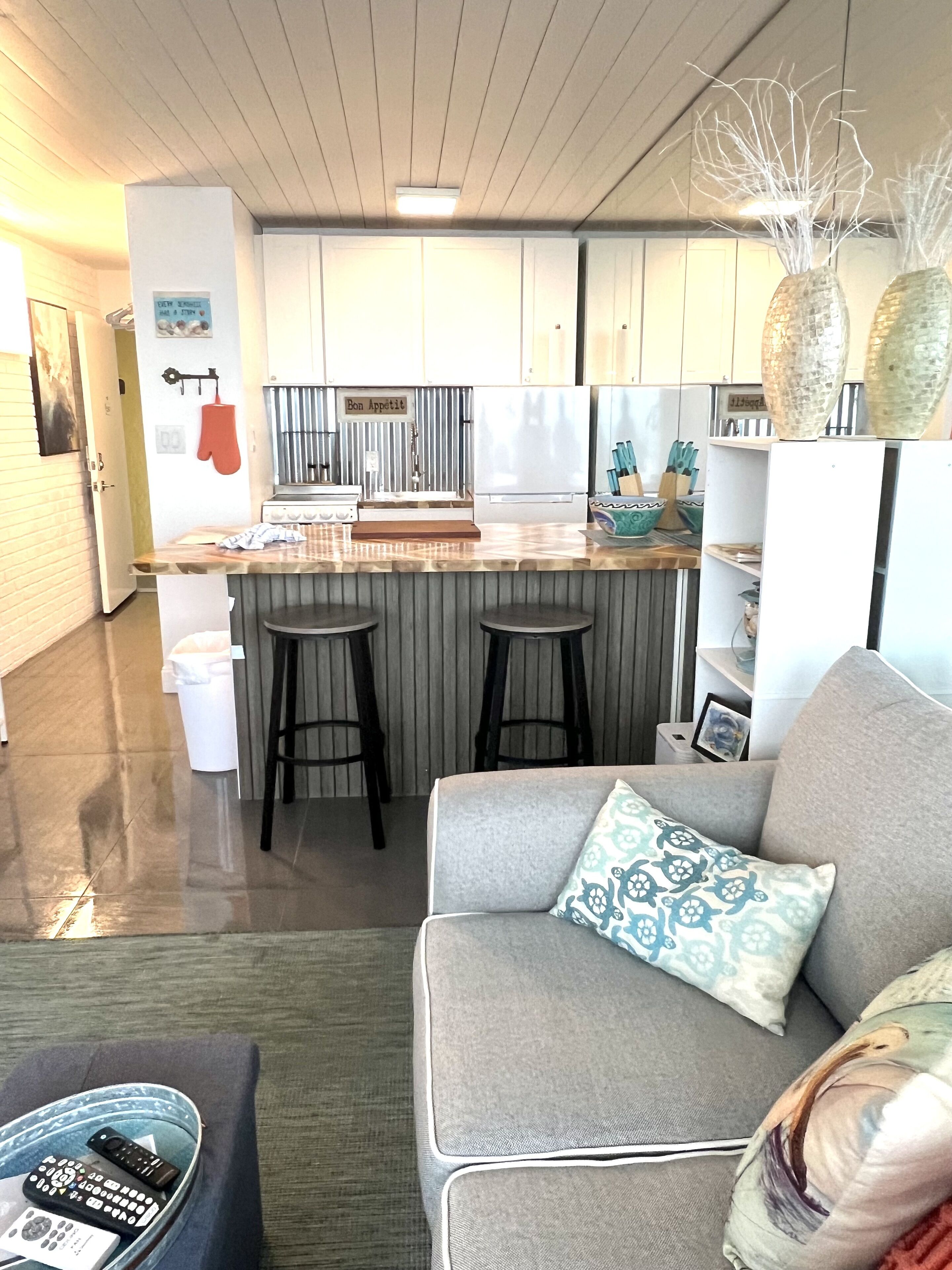 A modern kitchen with a central island, bar stools, and a dining area with a grey sofa and patterned cushions.