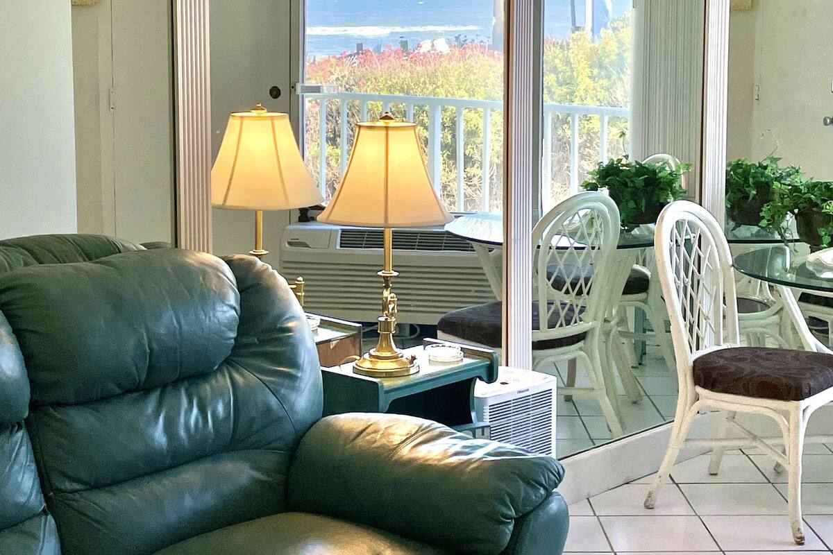 A room with a green leather sofa, two lamps, a table with chairs, and a view of the ocean through large windows.