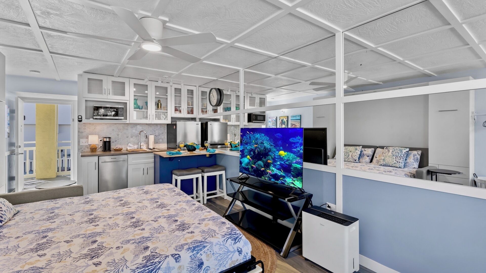 A compact studio apartment with a kitchenette, a bed, a desk with a computer, and a large aquarium.