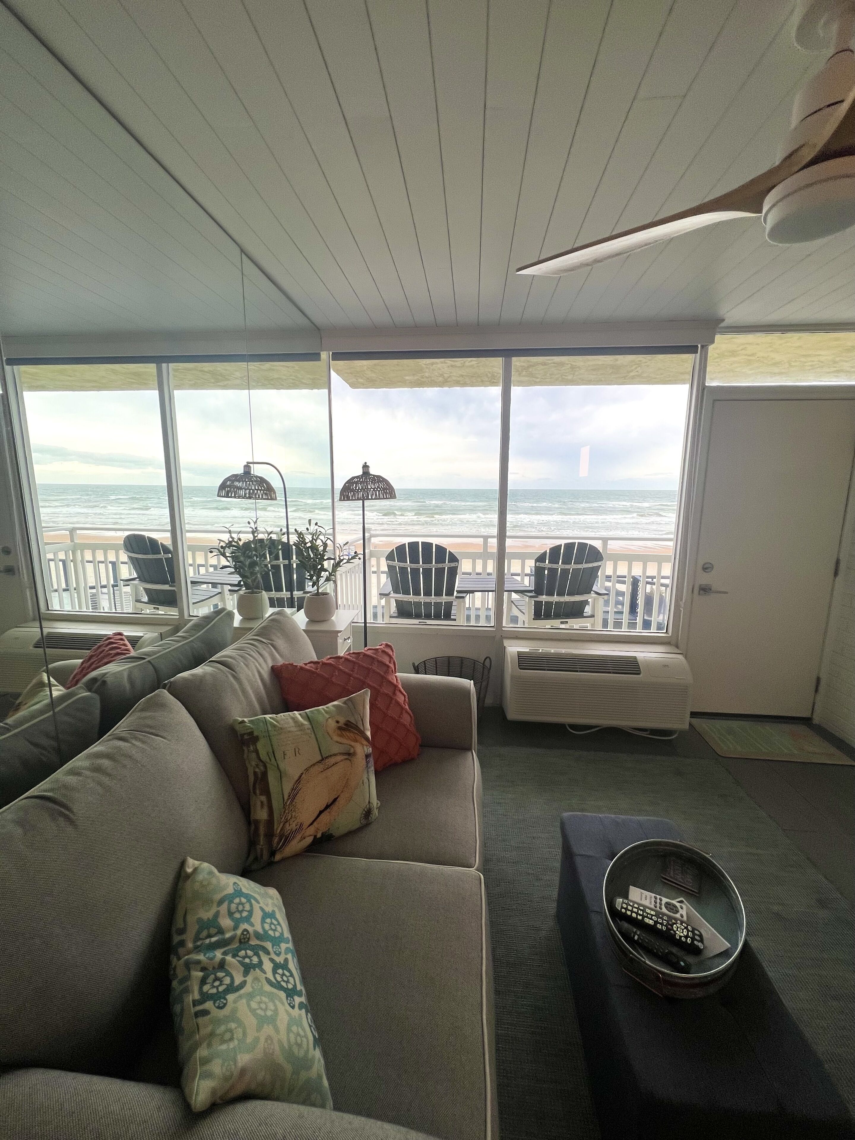 Una sala de estar con un ventanal grande con vista al océano, un sofá con cojines estampados, una mesita con una bandeja y un sillón junto a la ventana.