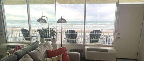A living room with a large window overlooking the ocean, a sofa with patterned cushions, a small table with a tray, and a chair by the window.